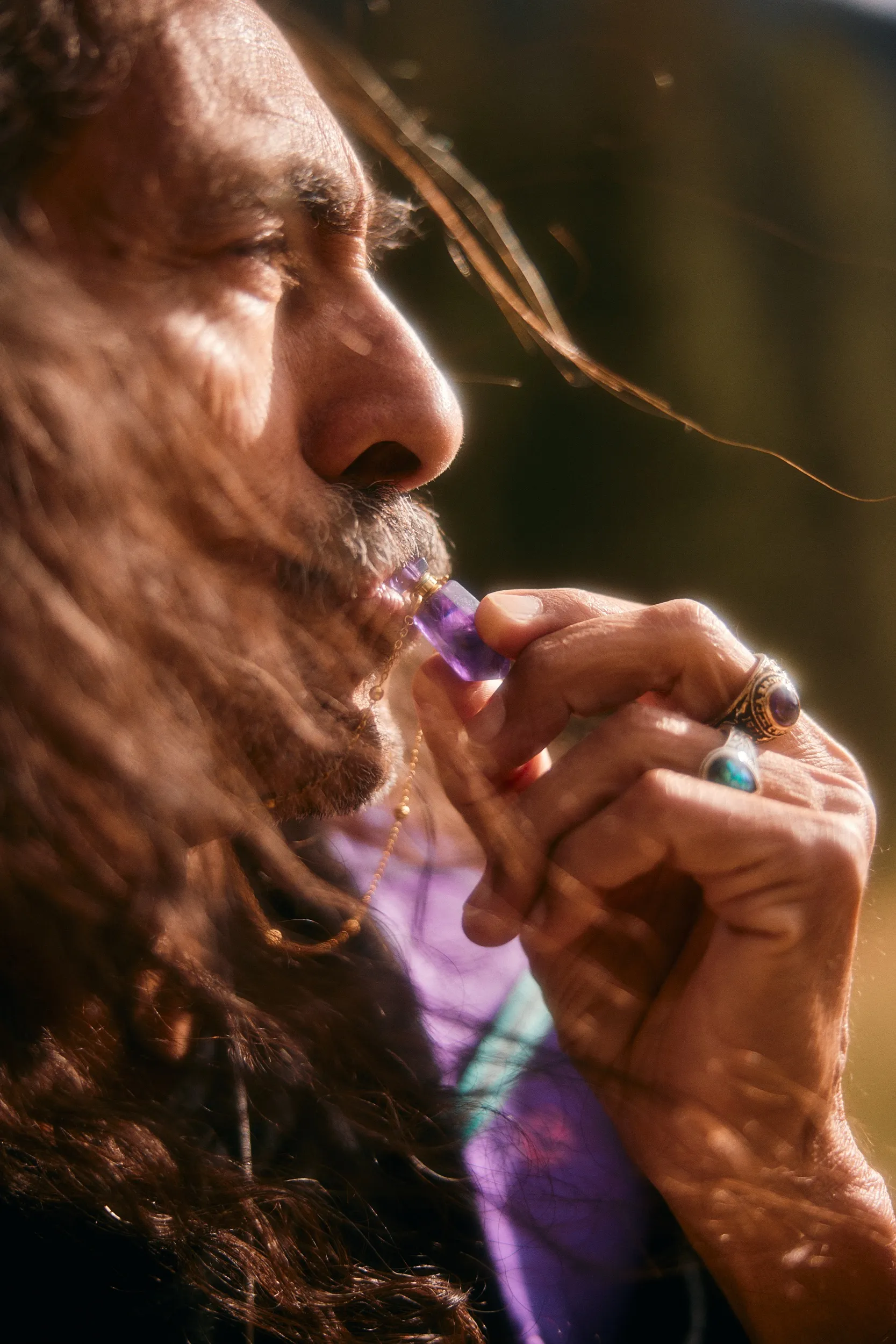 Profile portrait of a man with long hair and rings holding a purple crystal pendant to his lips